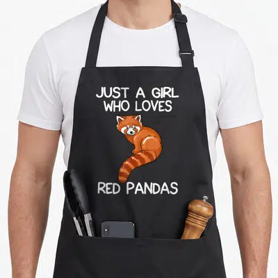 Nur panda apron - Cartoon red panda in orange and white between white text blocks on black — shown on a black cotton canvas apron against a white background