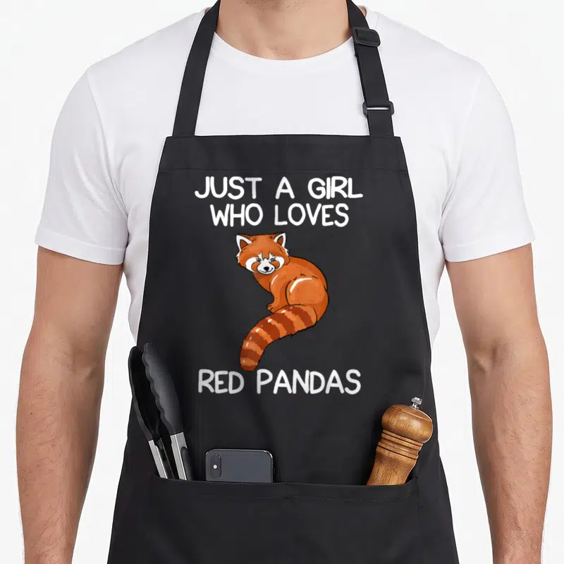 Nur panda apron - Cartoon red panda in orange and white between white text blocks on black — shown on a black cotton canvas apron against a white background