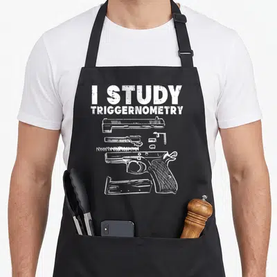Study pro apron - White text I Study Triggernometry above exploded handgun parts on distressed background — shown on a black cotton canvas apron against a white background