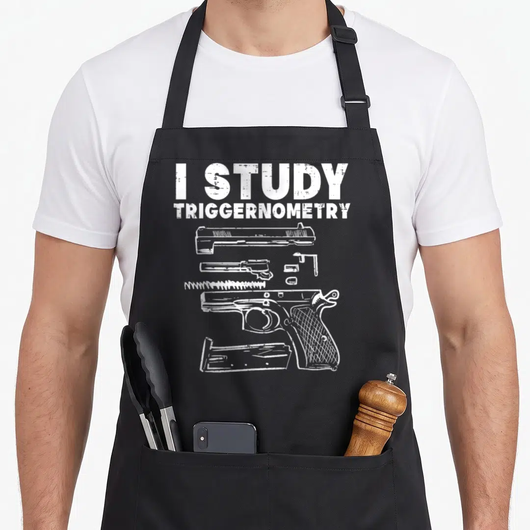 Study pro apron - White text I Study Triggernometry above exploded handgun parts on distressed background — shown on a black cotton canvas apron against a white background