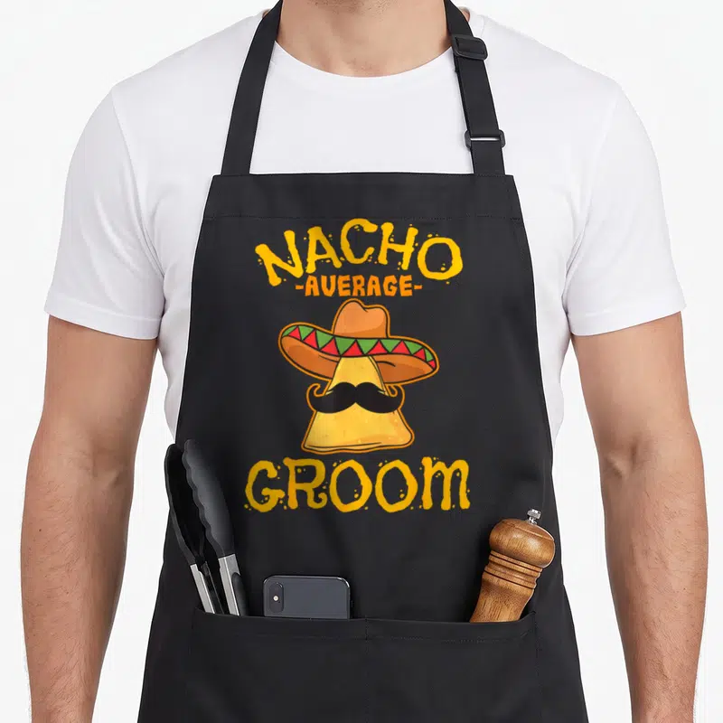 Groom mexican apron - Cartoon nacho chip wearing sombrero and mustache with NACHO AVERAGE GROOM text in yellow — shown on a black cotton canvas apron against a white background