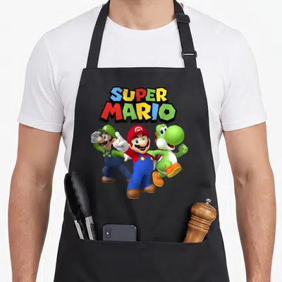 Cute shot apron - Super Mario logo in red blue green yellow above Mario Luigi and Yoshi standing in a group — shown on a black cotton canvas apron against a white background