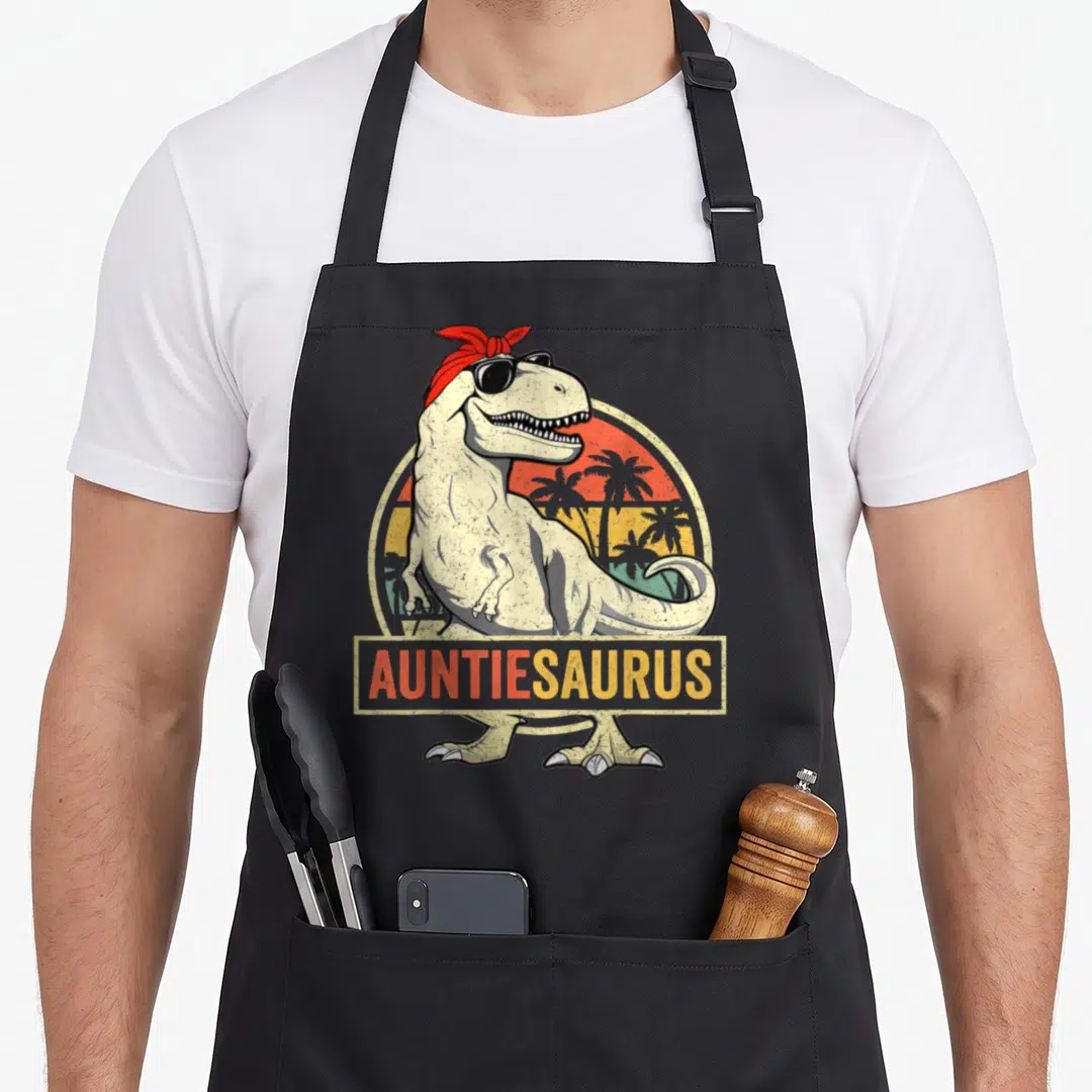 Saurus dinosaur apron - AUNTIESAURUS text over T-Rex with bandana and retro sunset in cream red yellow orange green — shown on a black cotton canvas apron against a white background