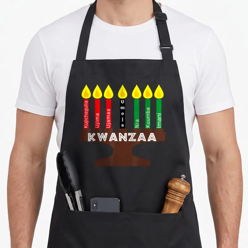 Kinara kwanzaa apron - Seven candles labeled with Kwanzaa principles atop a brown kinara holder with KWANZAA text — shown on a black cotton canvas apron against a white background