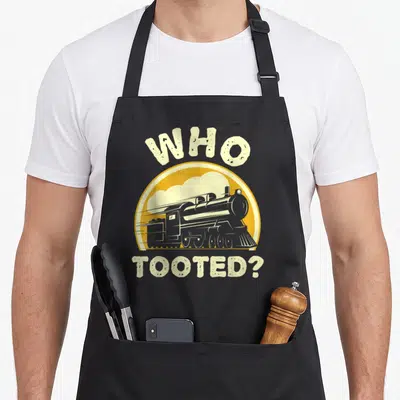 Tooted train locomotive railroad apron - Cartoon steam locomotive in yellow circle with WHO TOOTED? text in beige — shown on a black cotton canvas apron against a white background