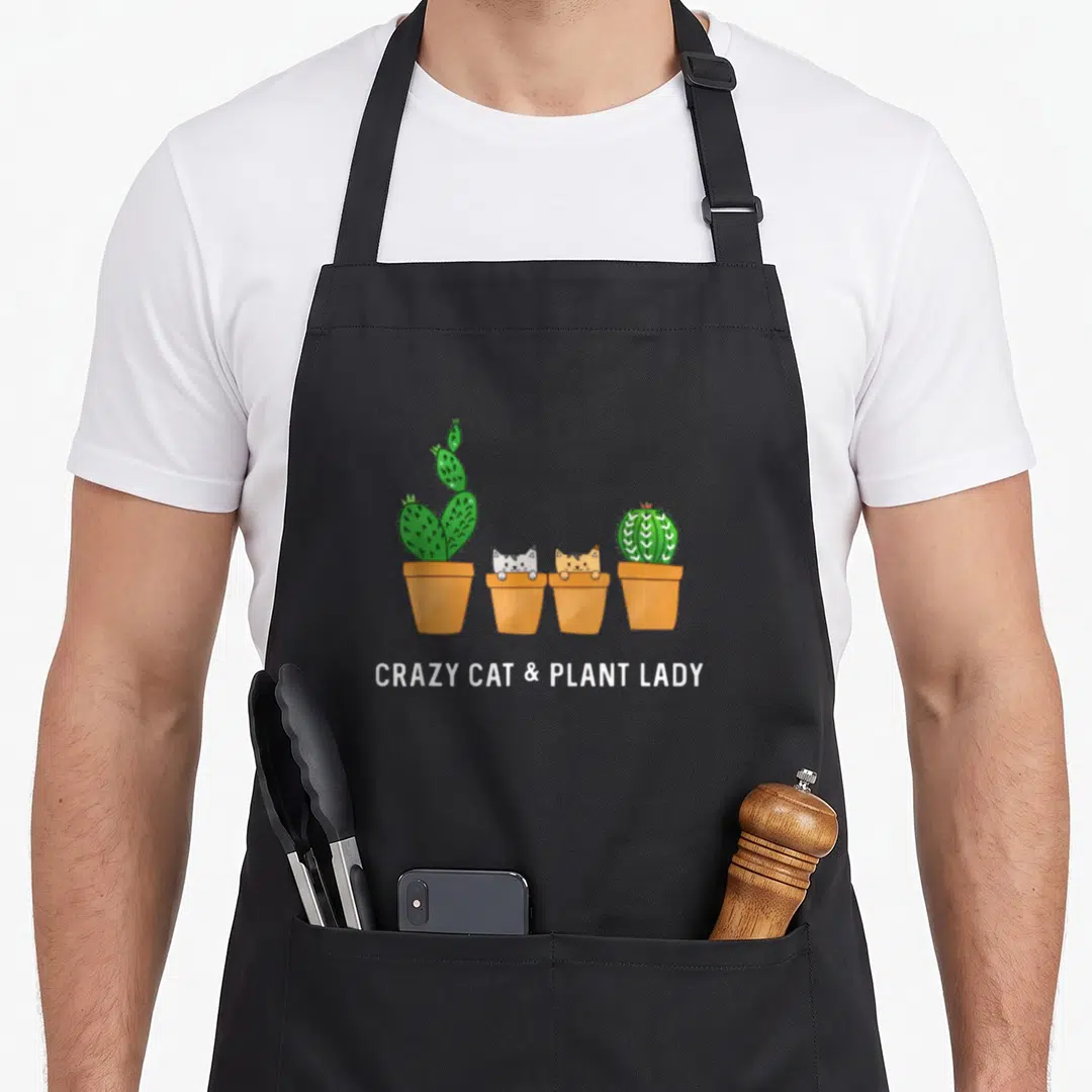 Cactus cat apron - Four terracotta pots holding green cacti and orange and white cats with white text below — shown on a black cotton canvas apron against a white background