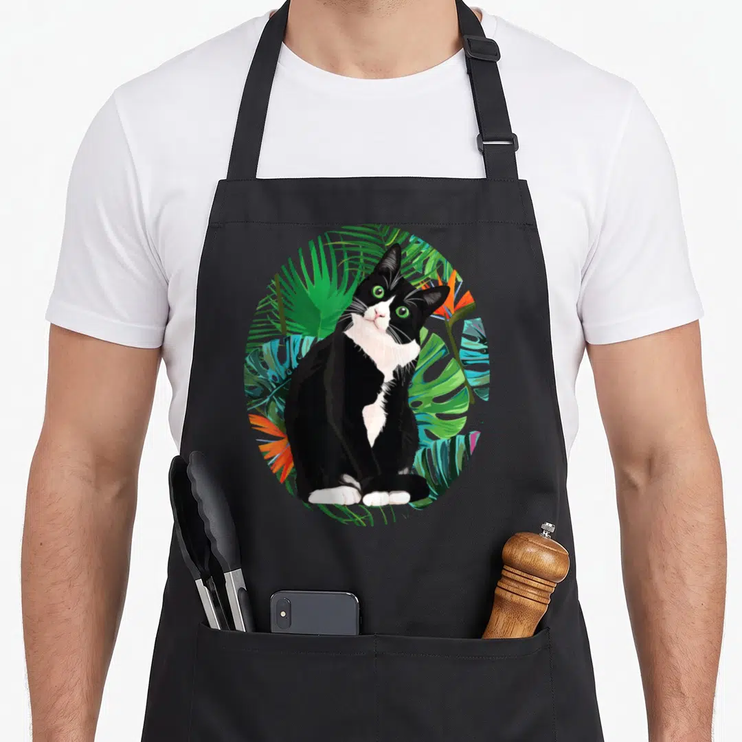 Hawaiian tuxedo apron - Cartoon tuxedo cat with green eyes among green palm leaves and orange flowers in a circle — shown on a black cotton canvas apron against a white background