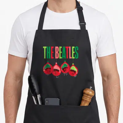 Ornament christmas santa apron - The Beatles text above four red ornaments with Santa hats and John Paul George Ringo — shown on a black cotton canvas apron against a white background