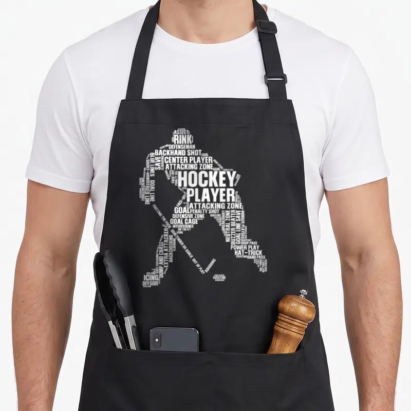 Cute player hockey ice apron - White typography silhouette of a hockey player formed by terms like RINK, GOAL — shown on a black cotton canvas apron against a white background