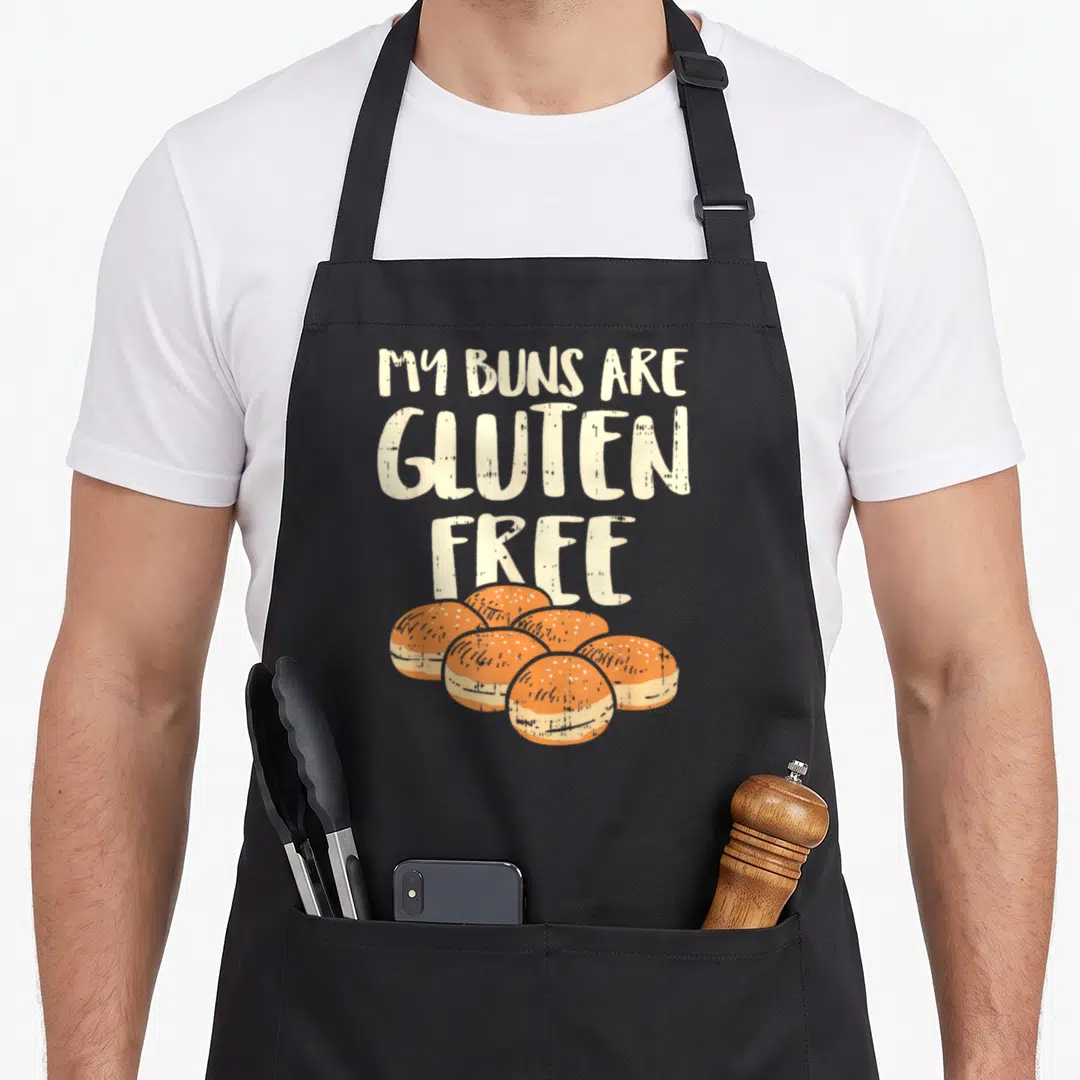 Gluten buns apron - Stacked cream text above six orange and cream cartoon burger buns — shown on a black cotton canvas apron against a white background