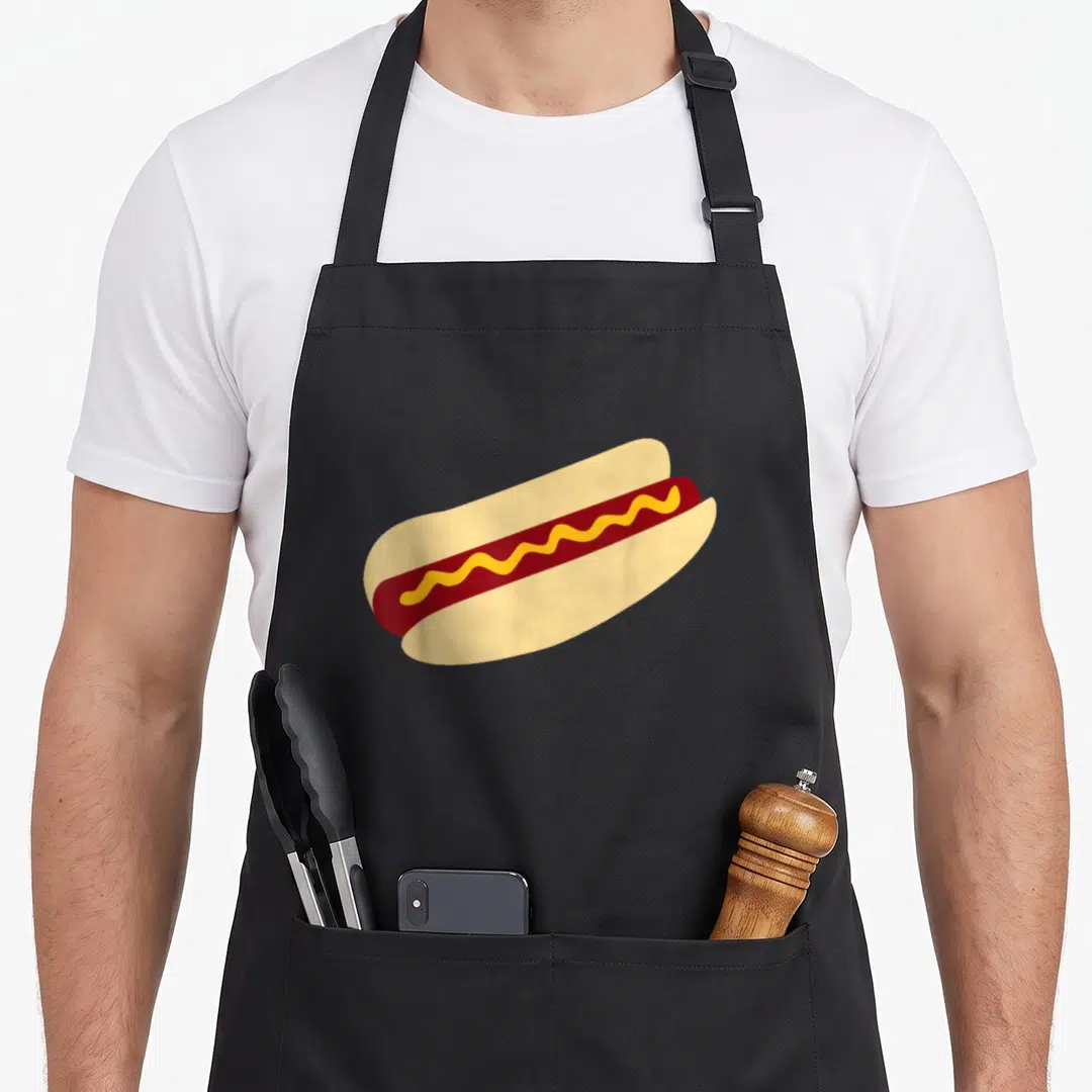 Cute hot dog apron - Single hot dog in tan bun with yellow mustard squiggle on dark red sausage — shown on a black cotton canvas apron against a white background
