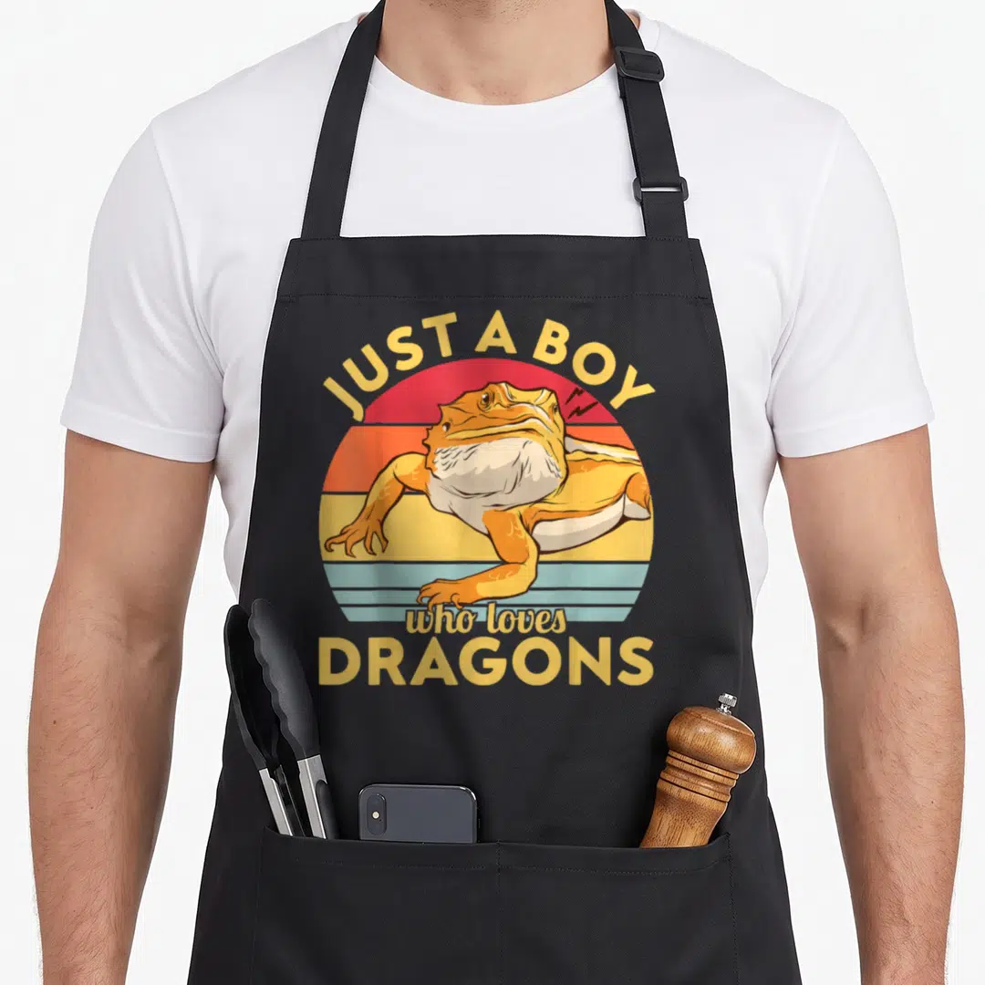 Cool bearded dragon apron - Retro toad in yellow orange red blue sunset circle with text JUST A BOY who loves DRAGONS — shown on a black cotton canvas apron against a white background