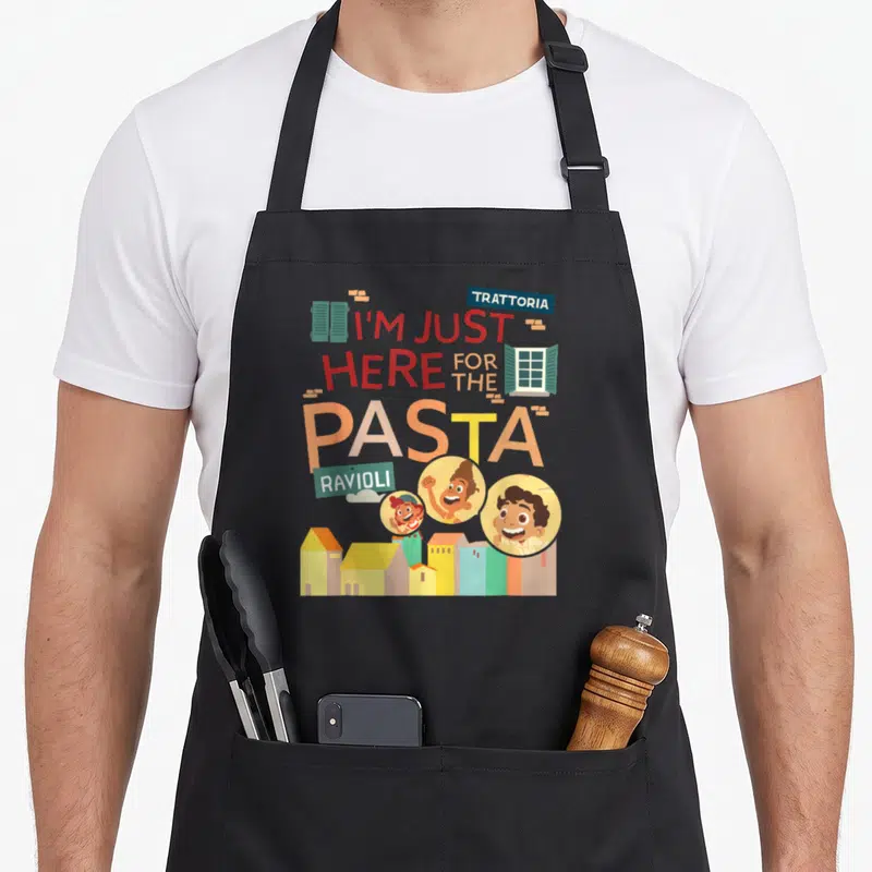 Luca pasta apron - Stacked text reading I'm just here for the pasta with cartoon faces and buildings below — shown on a black cotton canvas apron against a white background