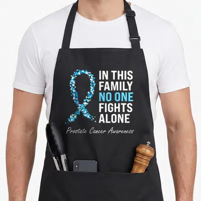 Light family aprons - Ribbon formed by small white and blue hearts next to stacked text about family fighting prostate — shown on a black cotton canvas apron against a white background