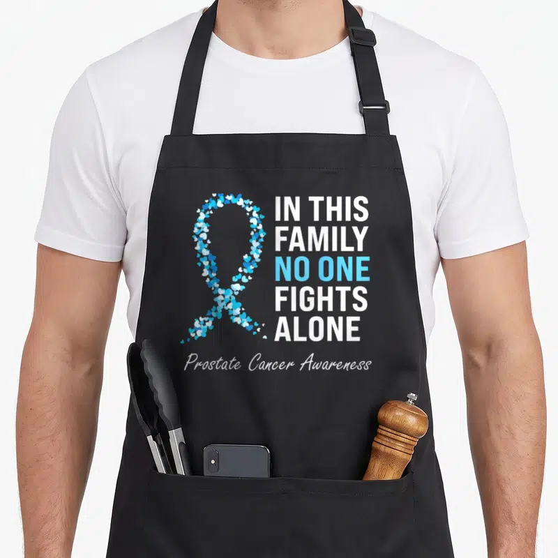 Light family aprons - Ribbon formed by small white and blue hearts next to stacked text about family fighting prostate — shown on a black cotton canvas apron against a white background