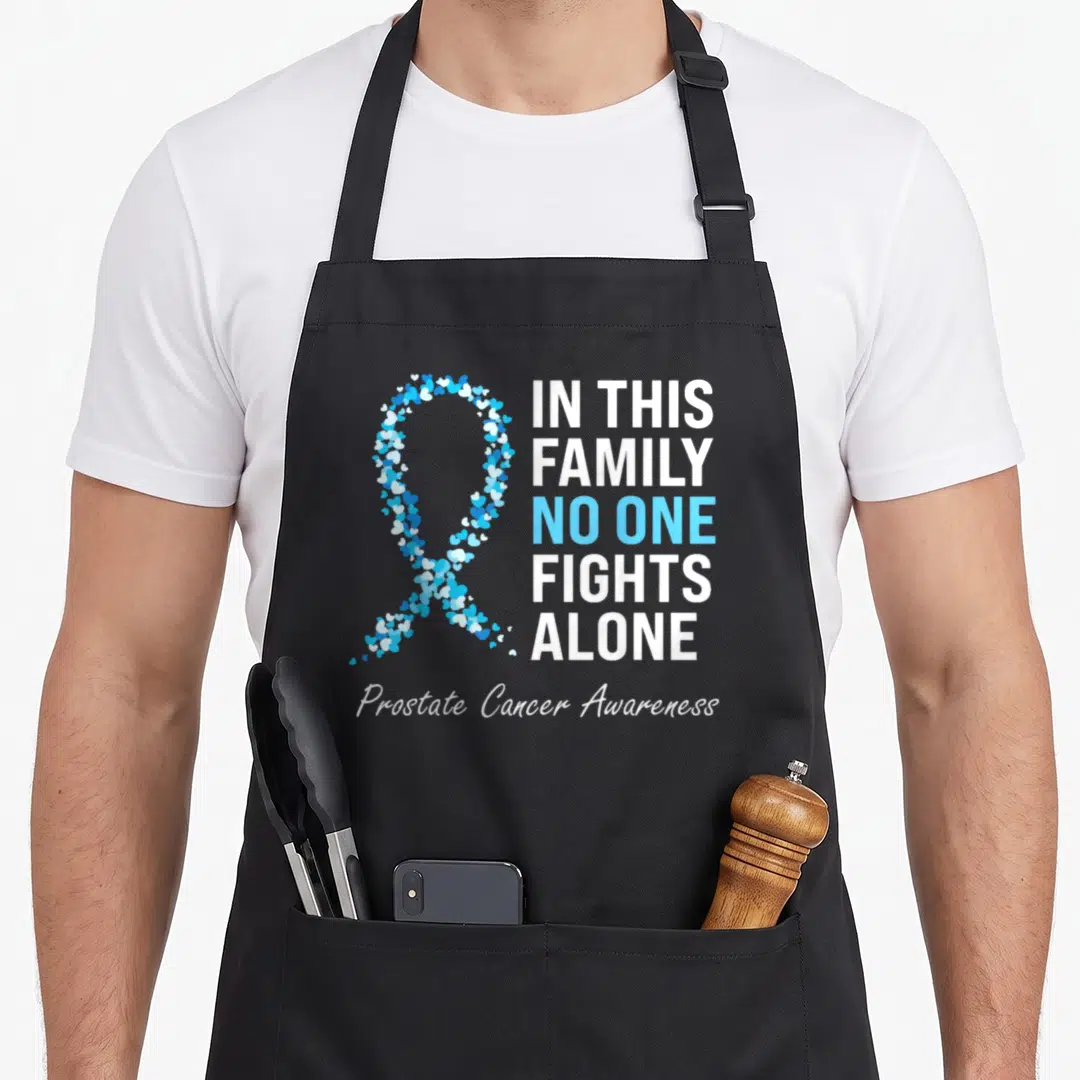 Light family aprons - Ribbon formed by small white and blue hearts next to stacked text about family fighting prostate — shown on a black cotton canvas apron against a white background