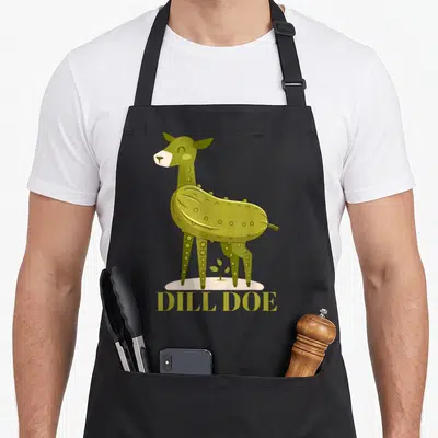 Doe pickle apron - Cartoon deer with pickle body, DILL DOE text, olive green and beige, small plant on beige ground — shown on a black cotton canvas apron against a white background