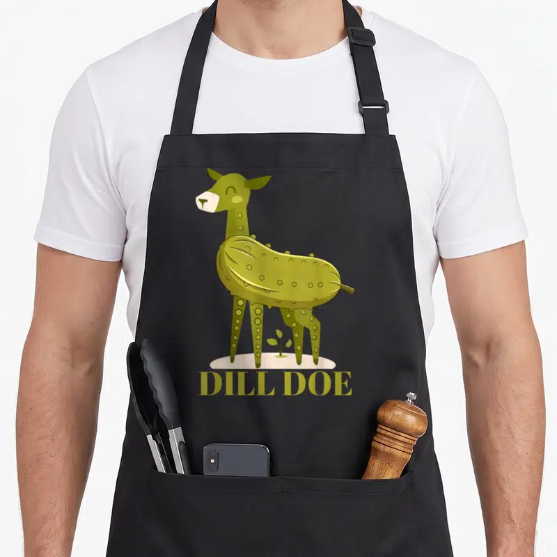 Doe pickle apron - Cartoon deer with pickle body, DILL DOE text, olive green and beige, small plant on beige ground — shown on a black cotton canvas apron against a white background