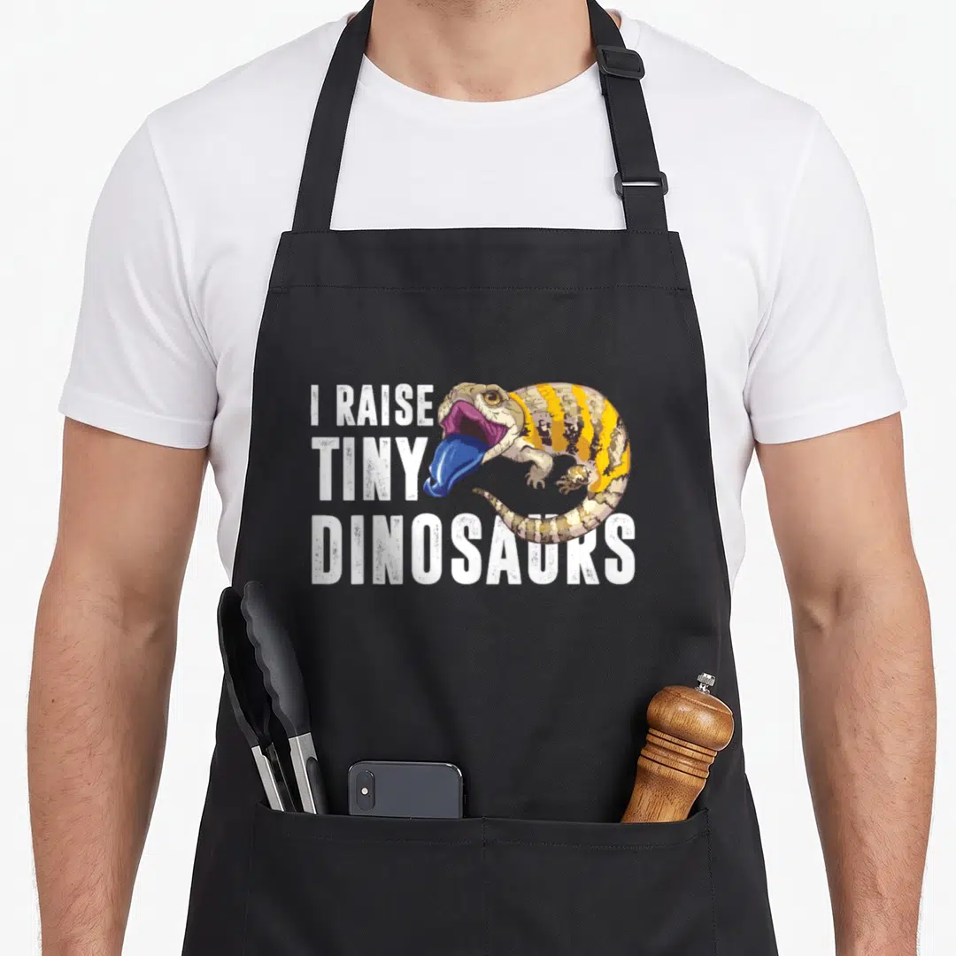 Blue dinosaur apron - Cartoon lizard with blue tongue overlaps white text reading I raise tiny dinosaurs — shown on a black cotton canvas apron against a white background