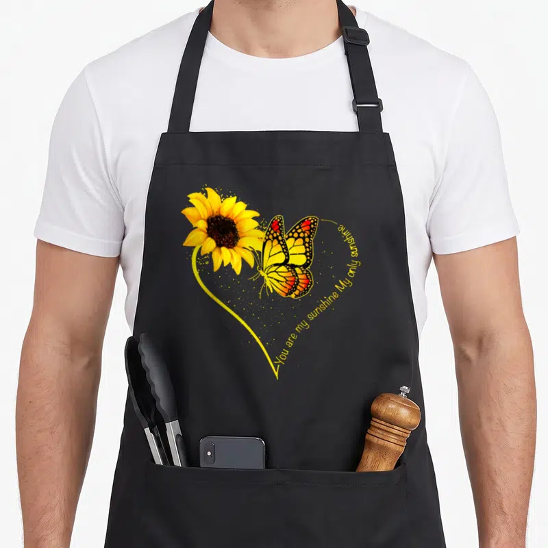 Butterfly sunflower apron - Realistic sunflower and butterfly with You Are My Sunshine text in a yellow heart outline — shown on a black cotton canvas apron against a white background
