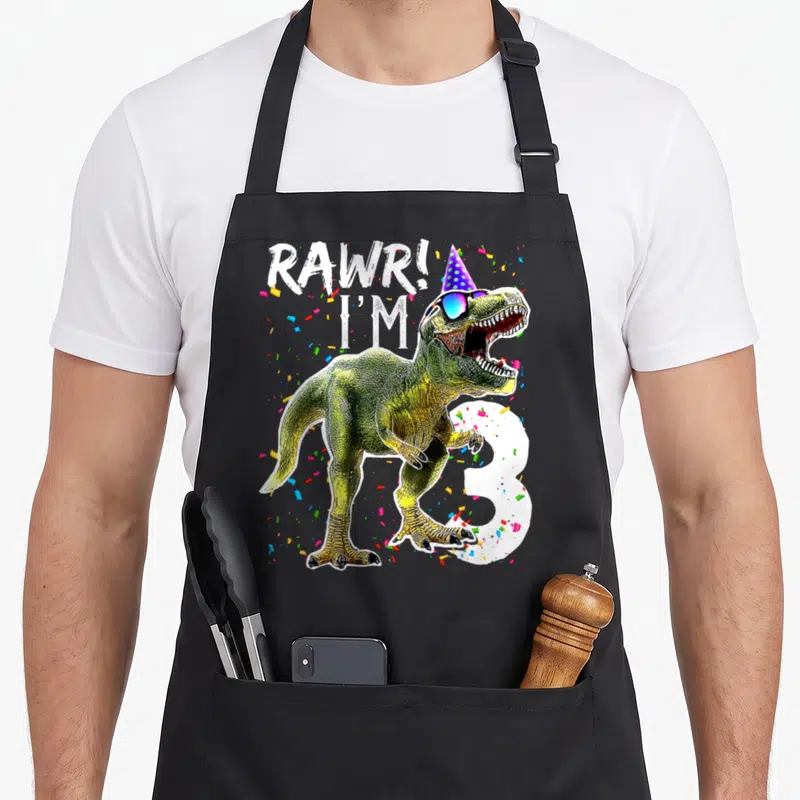 Rawr dinosaur apron - T-Rex wearing party hat and sunglasses with RAWR I'M 3 text and confetti — shown on a black cotton canvas apron against a white background