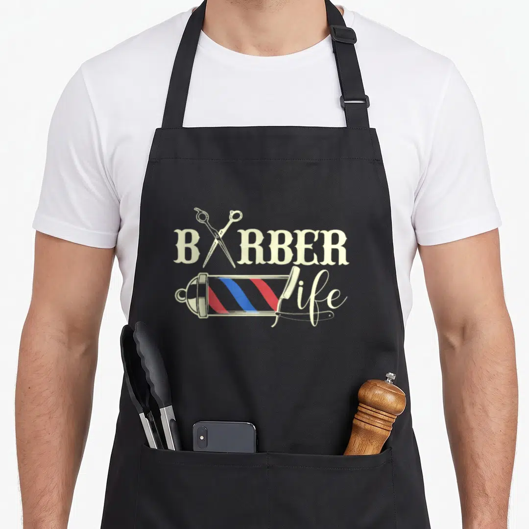 Barber husband life apron - Crossed scissors, barber pole with red blue stripes, and straight razor over text — shown on a black cotton canvas apron against a white background