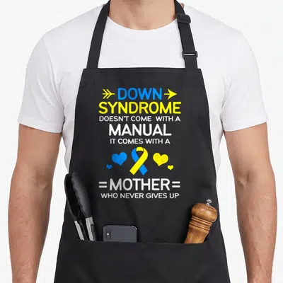Strong down syndrome awareness apron - Blue and yellow ribbon flanked by hearts with stacked text reading mother who — shown on a black cotton canvas apron against a white background