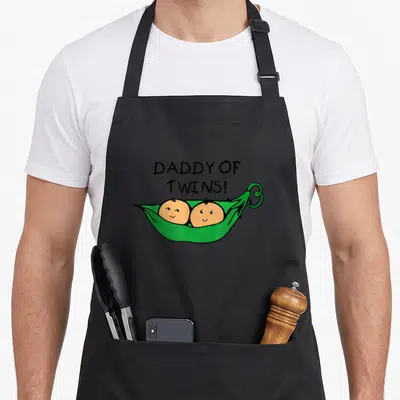 Peas daddy apron - DADDY OF TWINS text above cartoon green pea pod with two peach baby faces — shown on a black cotton canvas apron against a white background