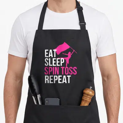 Eat color guard apron - Stacked EAT SLEEP SPIN TOSS REPEAT text with pink flag bearer silhouette — shown on a black cotton canvas apron against a white background