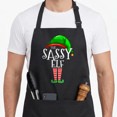 Sassy personalized family aprons - Green elf hat, white text, red striped socks, and curled green shoes on black fabric — shown on a black cotton canvas apron against a white background