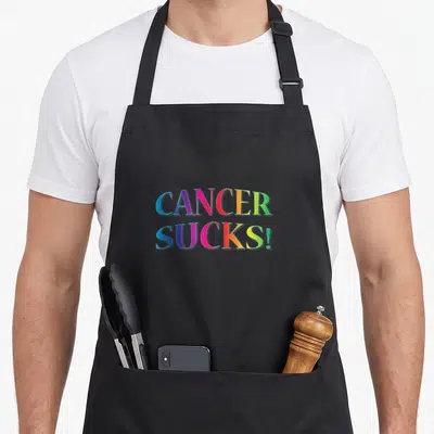 Cancer support your apron - CANCER SUCKS! text in rainbow gradient colors on black fabric — shown on a black cotton canvas apron against a white background