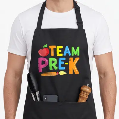Nursery cute teacher aprons - Colorful TEAM PRE-K text with red apple and orange crayon in cartoon style — shown on a black cotton canvas apron against a white background