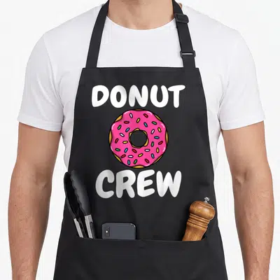 Doughnut sprinkle donut apron - White Donut Crew text surrounding a pink frosted cartoon donut with colorful sprinkles — shown on a black cotton canvas apron against a white background
