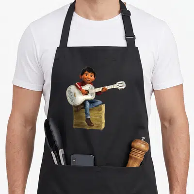Coco playing guitar apron - Boy in red hoodie playing white guitar on wooden crate — shown on a black cotton canvas apron against a white background