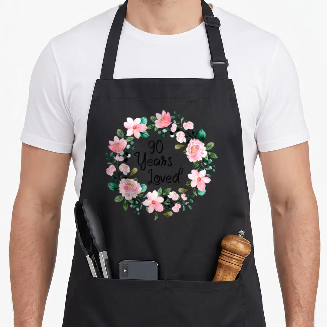 Loved floral aprons - Pink watercolor flower wreath surrounding black 90 Years Loved text — shown on a black cotton canvas apron against a white background