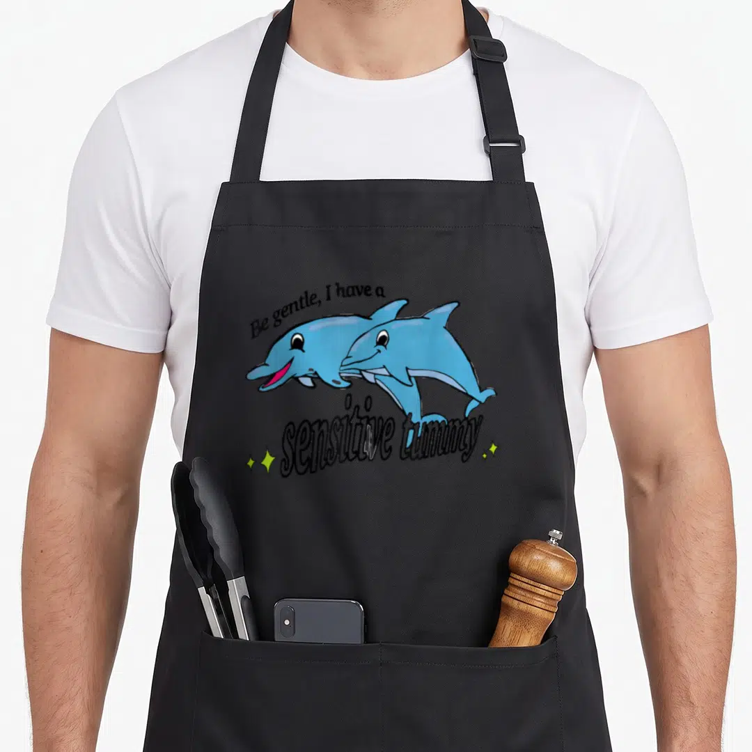 Sensitive gentle apron - Two blue cartoon dolphins jumping with text Be gentle I have a sensitive tummy flanked — shown on a black cotton canvas apron against a white background