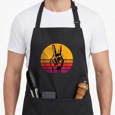 Inspired hippie apron - Black peace sign hand silhouette over yellow orange red purple striped sunset circle — shown on a black cotton canvas apron against a white background