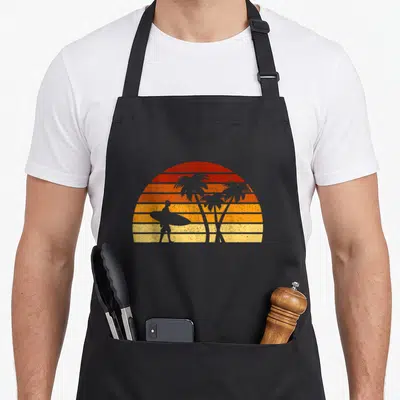 Sun surfer apron - Retro sunset with horizontal stripes, surfer silhouette, and three palm trees in red orange yellow — shown on a black cotton canvas apron against a white background