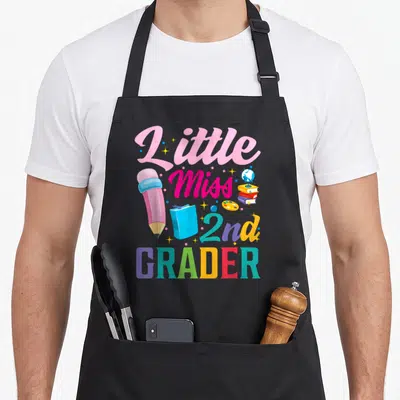 Miss 1st apron 2nd apron - Cartoon pink pencil, stacked books, and Little Miss 2nd GRADER text in pink, teal, yellow — shown on a black cotton canvas apron against a white background