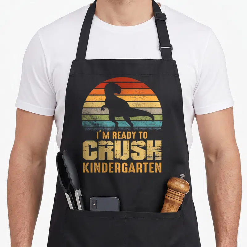 Crush kindergarten dinosaur apron - Grey dinosaur silhouette over striped sun with I'm Ready To Crush Kindergarten text — shown on a black cotton canvas apron against a white background