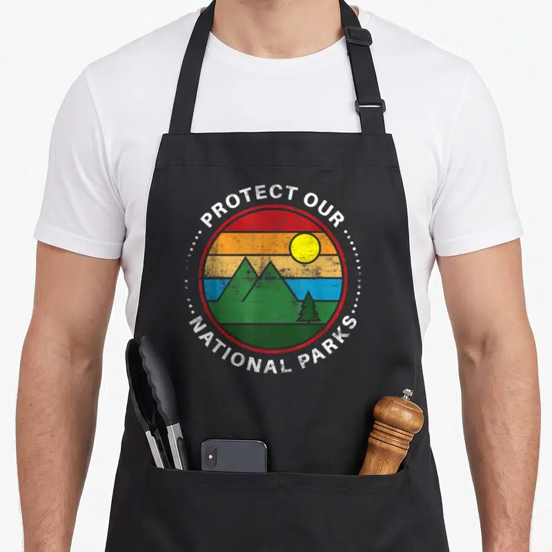 Yeoy modern apron - Retro circular badge with mountains, tree, sun and Protect Our National Parks text — shown on a black cotton canvas apron against a white background