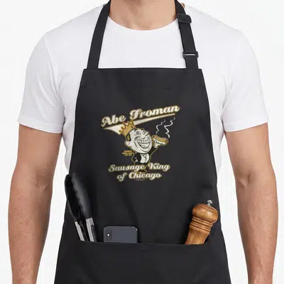 Bueller sausage apron - Cartoon man with crown holding hot dog, Abe Froman Sausage King text — shown on a black cotton canvas apron against a white background