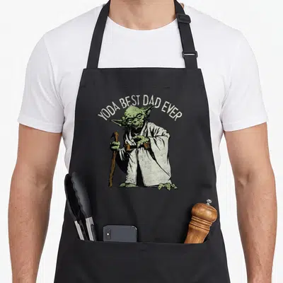Vintage yoda apron - Arched YODA BEST DAD EVER text above a green robed figure holding a brown cane — shown on a black cotton canvas apron against a white background