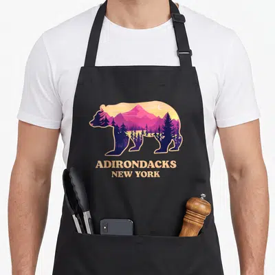 Adirondack mountain new york apron - Bear silhouette with purple mountains and trees above beige Adirondacks New York — shown on a black cotton canvas apron against a white background
