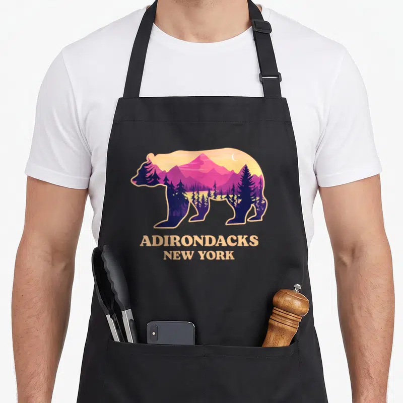 Adirondack mountain new york apron - Bear silhouette with purple mountains and trees above beige Adirondacks New York — shown on a black cotton canvas apron against a white background