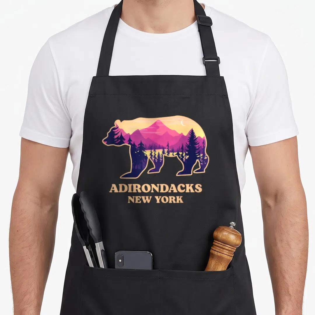 Adirondack mountain new york apron - Bear silhouette with purple mountains and trees above beige Adirondacks New York — shown on a black cotton canvas apron against a white background