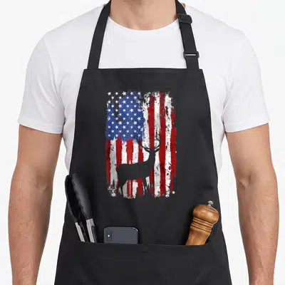 Deer hunter american flag apron - Grey deer silhouette standing before a distressed red white and blue American flag — shown on a black cotton canvas apron against a white background