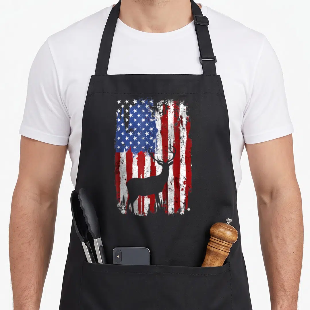 Deer hunter american flag apron - Grey deer silhouette standing before a distressed red white and blue American flag — shown on a black cotton canvas apron against a white background