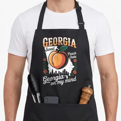 State southern aprons - Retro peach on white Georgia map with skyline and floral vines in orange and green — shown on a black cotton canvas apron against a white background