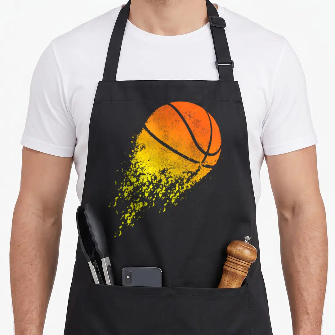 Basketball sports fan apron - Orange basketball with yellow splattered tail and distressed texture on grey background — shown on a black cotton canvas apron against a white background